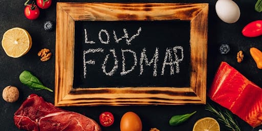 Quadro negrito escrito em branco "LOW FODMAR" com algumas carnes, legume s e verduras espalhadas na mesa